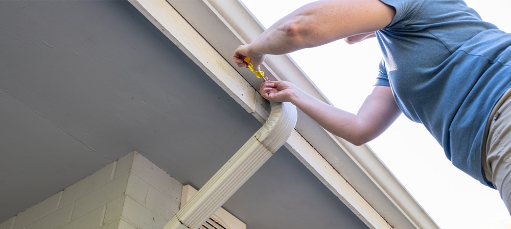 middle aged white woman working overhead on a ladder with a screwdriver removing screw from metal downspout bracket at roof gutter