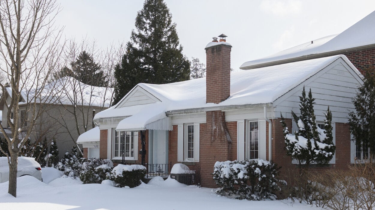 suburban house stands covered in snow with icicles hanging from the roof, while the sunlight casts shadows on the white winter landscape