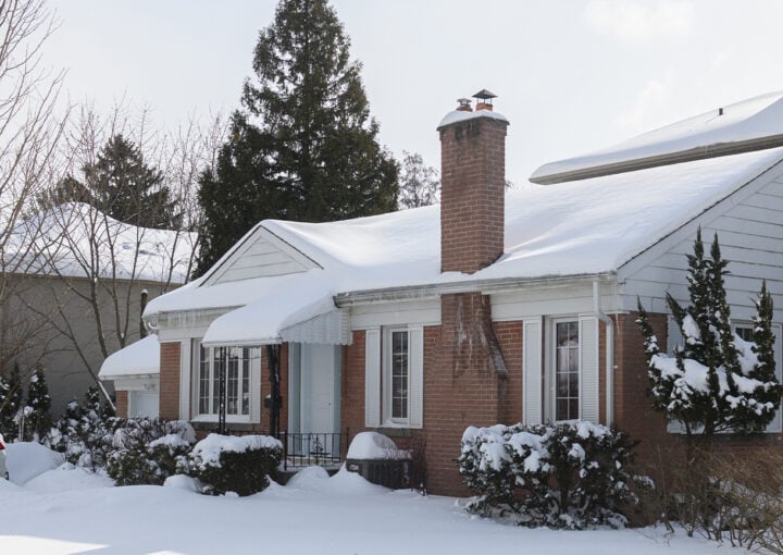 suburban house stands covered in snow with icicles hanging from the roof, while the sunlight casts shadows on the white winter landscape
