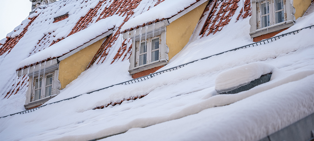 The scene showcases a charming rooftop blanketed in snow, with vibrant yellow panels visible under thick layers of white. Icicles dangle elegantly from the edges, capturing the essence of winter.
