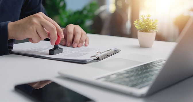 Individual in an office setting applies a stamp to a document contract legal agreement, signifying approval or verification certification permit visa next to an open laptop.