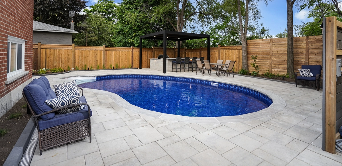 interlocking surface around pool, open kitchen with sitting space and fence with landscaping in home backyard