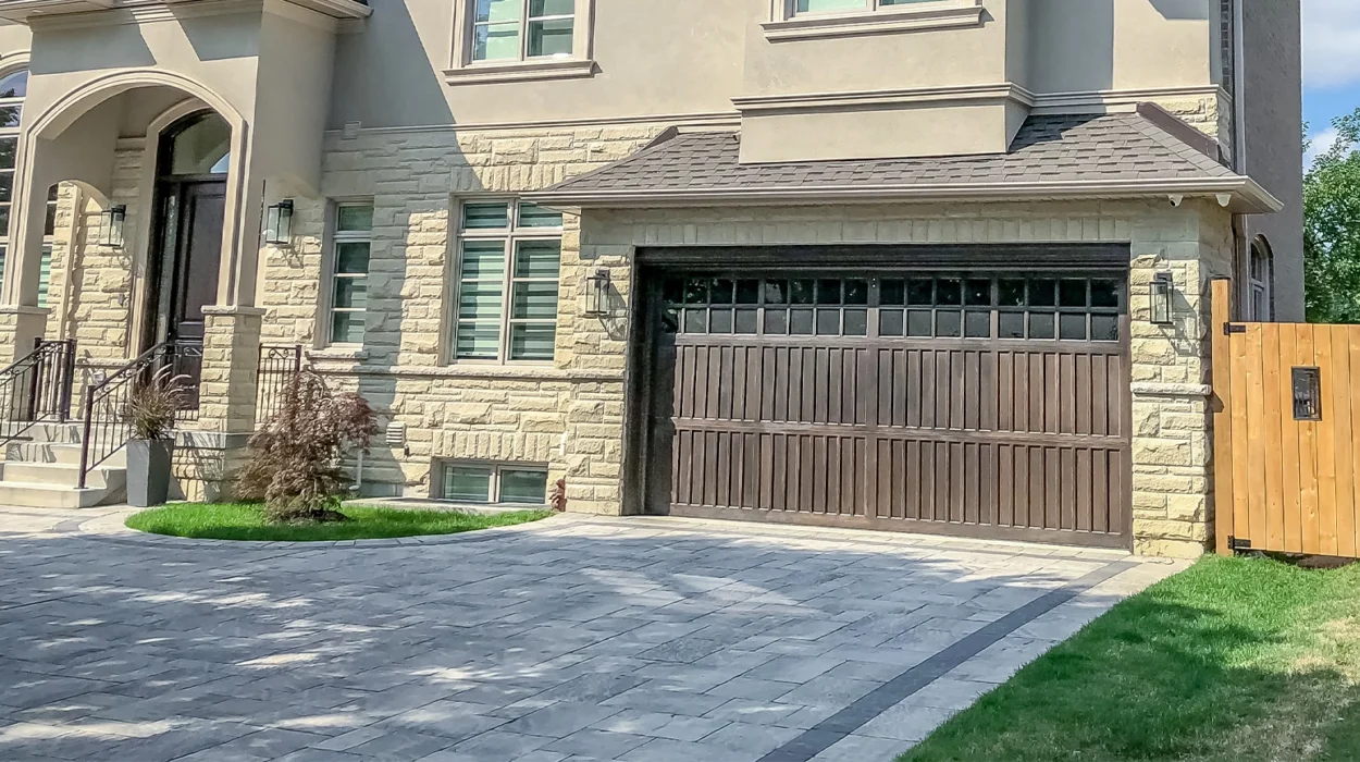 A beautiful large modern custom executive residential house with grey stone and brick, with gray shingles and grey trim, patterned interlock walkway and driveway, and double garage doors.