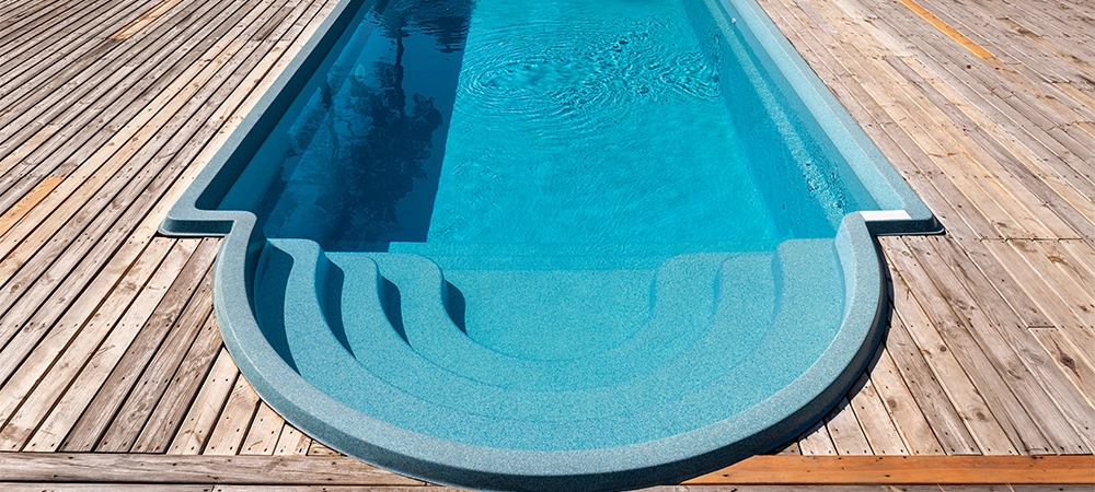 New modern fiberglass plastic swimming pool entrance step with clean fresh refreshing blue water on bright hot summer day at yard or resort hotel spa area. Wooden flooring deck of teak or larch board