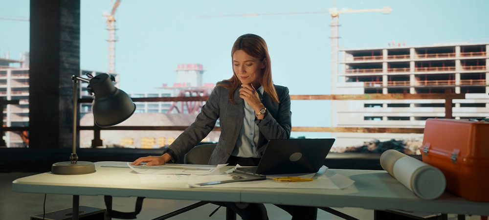 Confident businesswoman examining building plans at desk with laptop, illuminated by lamp, with large windows showcasing a bustling construction site in the background