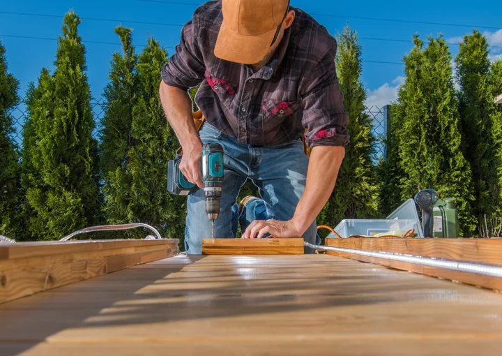 A carpenter is using a drill to secure wooden planks while constructing a deck outdoors on a sunny day. Lush trees are visible in the background.
