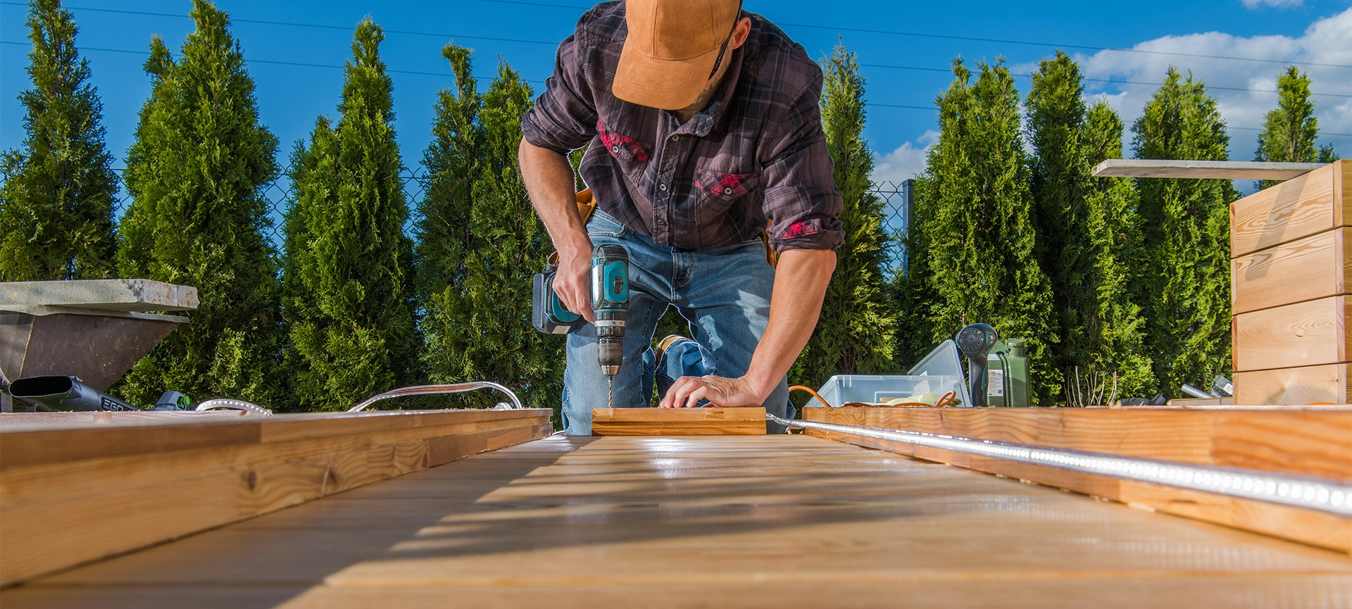 A carpenter is using a drill to secure wooden planks while constructing a deck outdoors on a sunny day. Lush trees are visible in the background.
