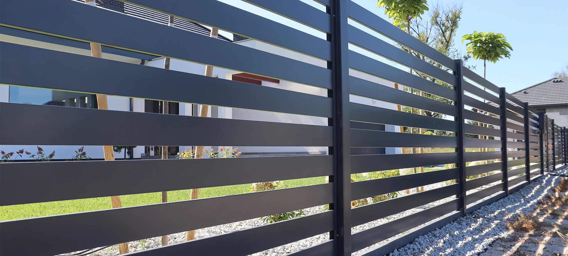 A sleek horizontal slat fence in a dark gray finish lines the perimeter of a modern home, complemented by decorative gravel and neatly planted ornamental trees under a clear blue sky.