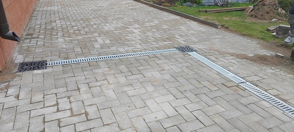 Workers are laying stone pavement in a yard. They are placing drainage channels to manage water flow. Bags are seen in the background, along with greenery and simple tools.