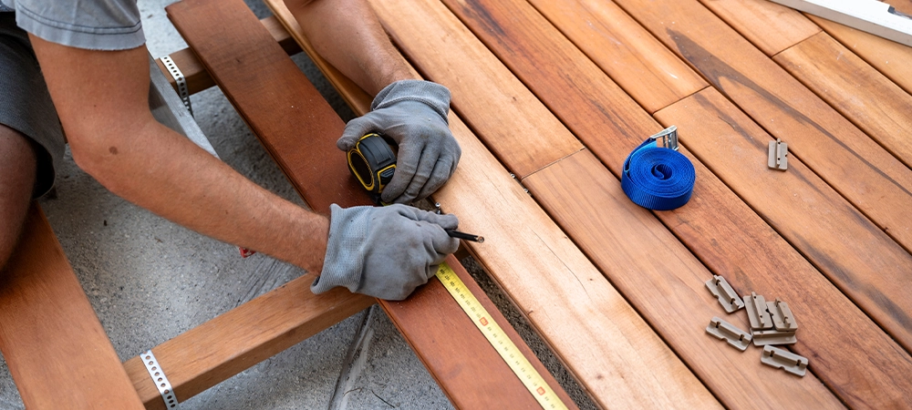 A craftsman wearing gloves measures and installs wooden decking, using tools like a tape measure and drill while ensuring precision in construction.