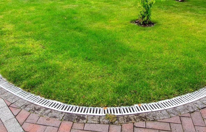 curved iron grid drainage system in the backyard by the green lawn with tree and footpath of red paving slabs.