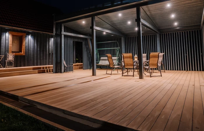 A modern wooden terrace at night with outdoor seating, a pergola, and warm lighting. A cozy ambiance with a covered dining area, a swing, and decorative lanterns.