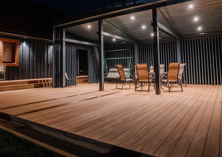 A modern wooden terrace at night with outdoor seating, a pergola, and warm lighting. A cozy ambiance with a covered dining area, a swing, and decorative lanterns.